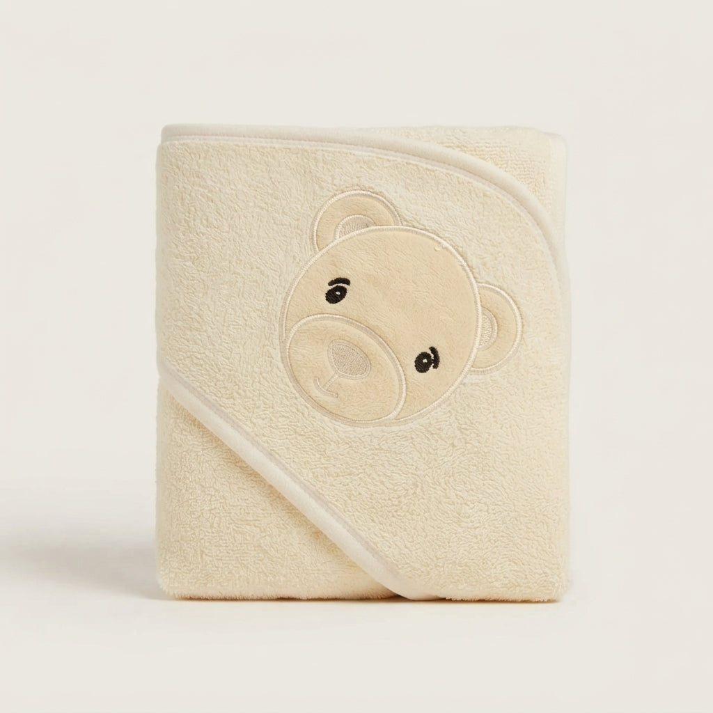 A folded cream-colored hooded baby towel with an embroidered teddy bear face on the front, set against a plain off-white background.