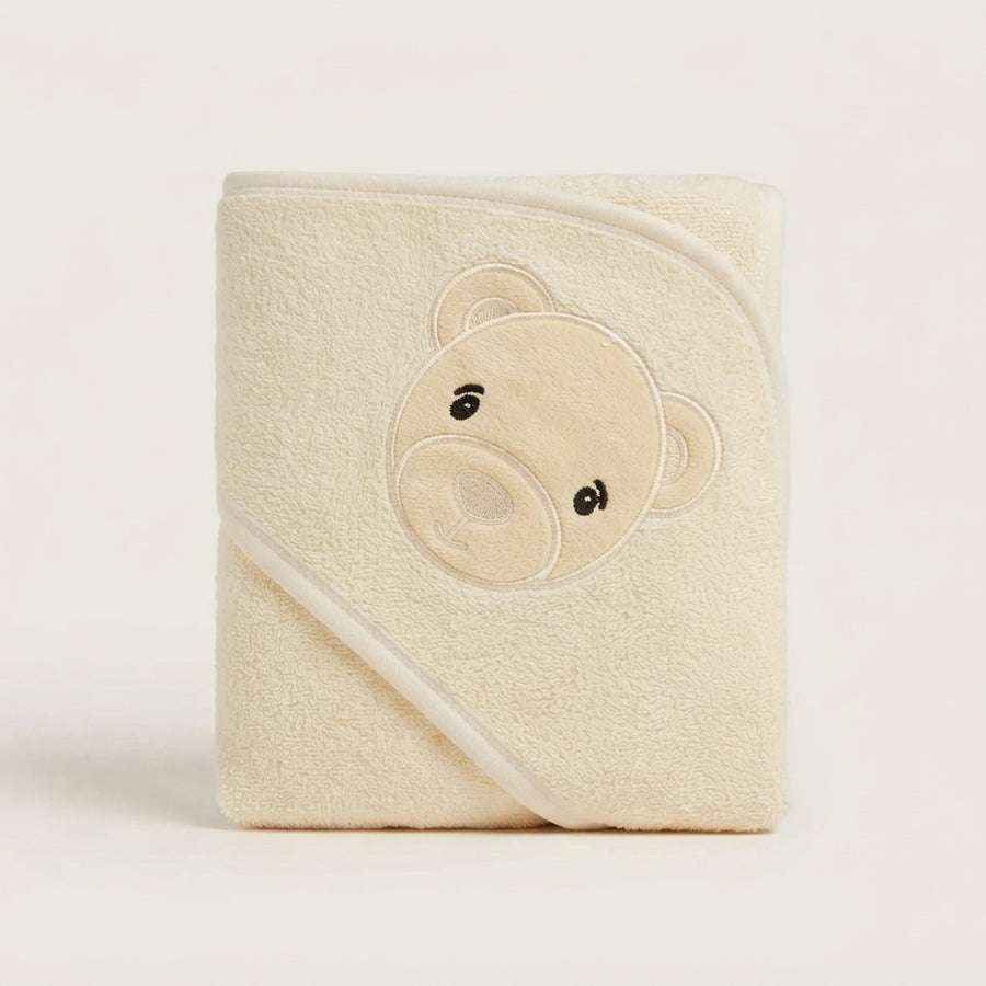 A folded cream-colored hooded baby towel with an embroidered teddy bear face on the front, set against a plain off-white background.