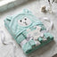 A folded mint green hooded towel with a sheep face design on the hood, tied with a white ribbon and tag that reads 'IORA GARMENTS'. The towel is styled on a white marble surface with a small decorative vase in the background.